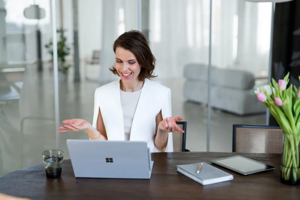 Sales Trainer Franziska Zimmermann sitzt an einem Tisch während eines Onlinemeetings und lacht in die Kamera des Laptops vor sich