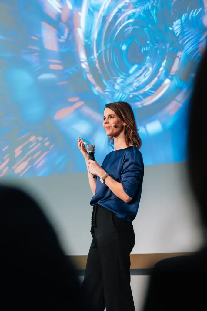 Franziska Zimmermann auf der Bühne mit einem Mikrofon in der Hand und in einer blauen Bluse während einer Wirtschaftsmoderation